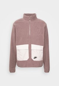 Pull en polaire mauve avec col montant, zip demi-longue à l'avant et deux poches blanches surdimensionnées. Présente un petit logo Nike noir sur la poche.
