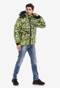 Schwarze und neongelbe Steppjacke mit auffälligem Logo-Print, Reißverschluss und Kapuze; kombiniert mit hellblauen Jeans und schwarzen Sneakers.