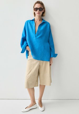 Frau trägt ein übergroßes, knallblaues Langarmshirt, beige weite knielange Shorts, weiße Schuhe und dunkle Sonnenbrille und steht vor einer weißen Wand.