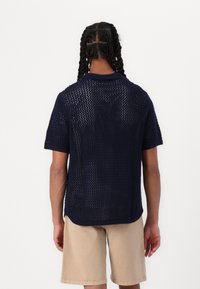 Polo de punto de color navy con mangas cortas, patrón de malla texturizada y cuello redondeado, combinado con pantalones cortos beige. La parte trasera tiene un corte holgado.