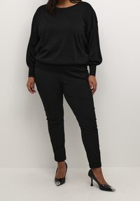 Gros pull noir oversize avec encolure ronde, manches longues et poignets côtelés. Associé à un pantalon noir ajusté et des escarpins pointus en cuir verni.