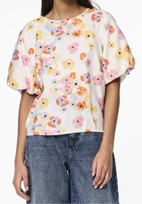 Donna che indossa una blusa bianca a maniche corte con maniche a sbuffo e stampato floreale multicolore, abbinata a jeans di denim blu.