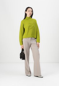 Heller grüner Rippstrickpullover mit hohem Kragen und erhabenem Muster, kombiniert mit beigen lässigen Hosen und einer schwarzen Handtasche.