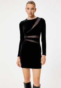 Minidress in velluto nero con maniche lunghe, caratterizzato da dettagli in rete trasparente e una silhouette aderente. Texture liscia con dettagli di design che esaltano le curve.