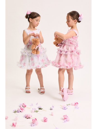 Δύο κορίτσια με φλοράλ φορέματα κρατούν αρκουδάκια, στέκονται πάνω σε λευκό δάπεδο σκορπισμένο με ροζ και μωβ λουλούδια.