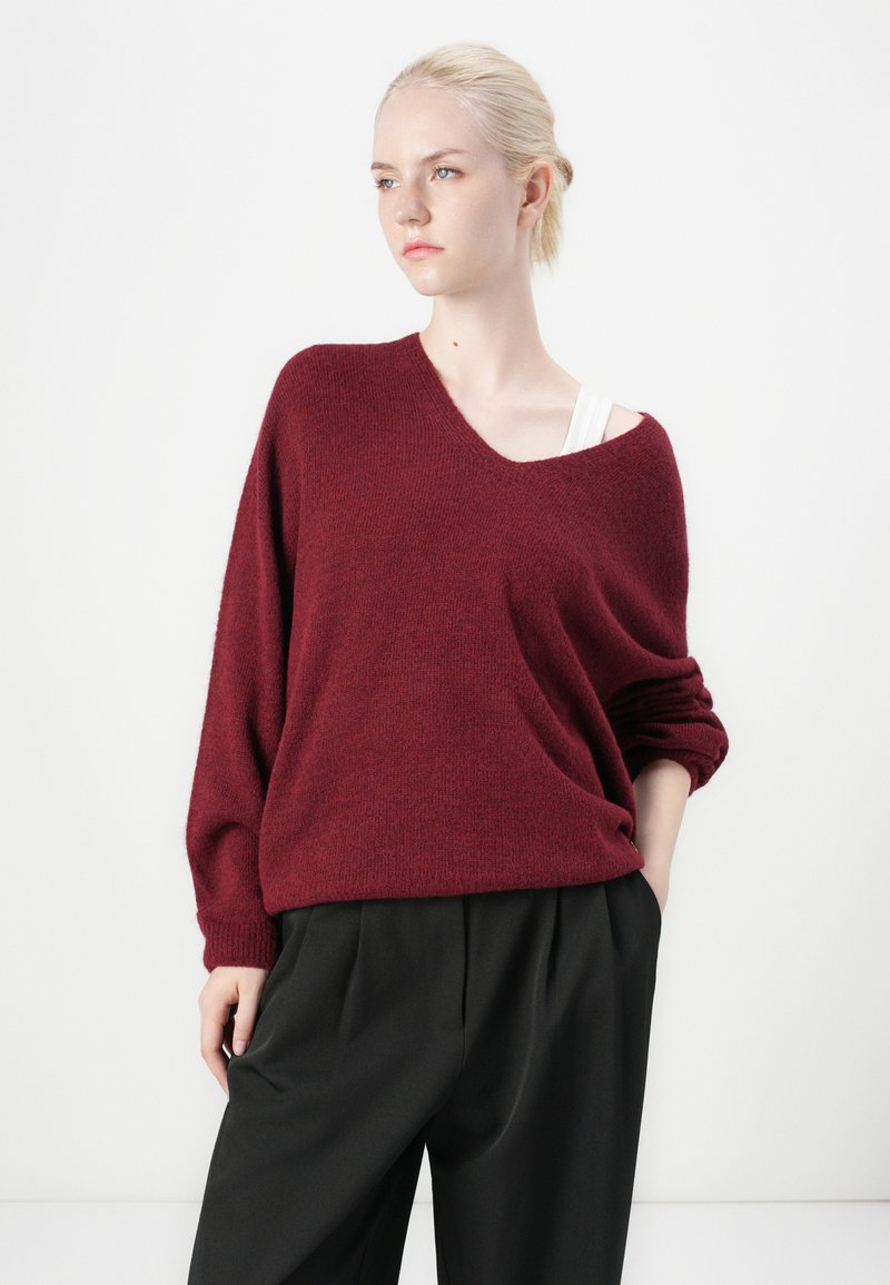 Burgunderfarbener Oversized-Pullover aus weichem Material mit tiefem V-Ausschnitt und umgeschlagenen Ärmeln, kombiniert mit hoch geschnittenen schwarzen Hosen.