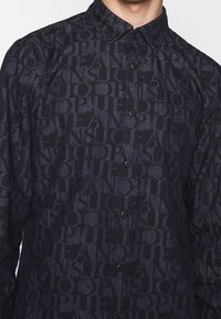 Camicia button-up blu scuro con un motivo di testo ripetuto in azzurro più chiaro, con colletto e bottoni neri. Tessuto morbido e testurizzato.