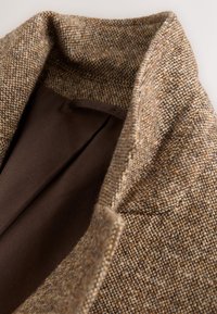 Col col en tweed marron avec un motif texturé, révélant une doublure intérieure marron foncé. Mise au point sur l'apparence tissée du tissu et la forme du col.