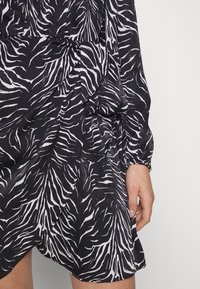 Šaty s potiskem zebry v černé a bílé barvě, vyrobené z měkkého materiálu, s dlouhými rukávy a asymetrickým lemem. Design zahrnuje nařasené detaily v oblasti pasu.