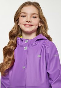 Lila vízálló dzseki kapucnival, ezüst színű patentgombokkal, egy kis esernyő logóval és élénk sárga béléssel.