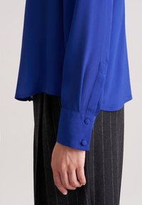 Langärmlige blaue Bluse aus glattem Stoff mit geradem Saum und geknöpften Manschetten, kombiniert mit strukturierten, dunklen Nadelstreifenhosen.