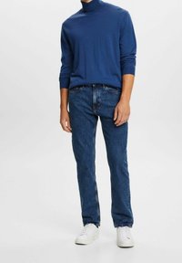 Hombre con un suéter de cuello alto azul, jeans rectos azules y zapatillas blancas de pie contra un fondo claro liso.