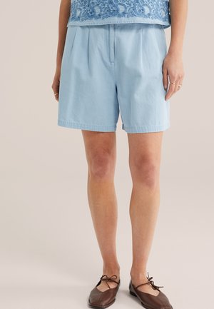 Šviesiai mėlyni klostuoti šortai, dėvimi su rudos spalvos bateliais su raišteliais, derinami su mėlynu siuvinėtu viršutiniu drabužiu.