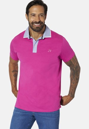 Man with beard and tattoos smiling, wearing pink polo shirt with striped collar and blue jeans, hands resting on hips.
