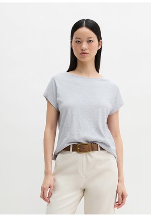 Junge Frau mit langen dunklen Haaren, die ein hellgraues kurzärmeliges Oberteil, beige Hosen und einen braunen Gürtel trägt, vor einem schlichten Hintergrund stehend.