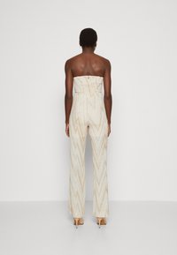 Ärmelloser Jumpsuit in Creme mit goldenen Zickzackmustern, ausgestattet mit einem taillierten Oberteil und weiten Hosenbeinen. Weiche Textur und zwei Knöpfe auf der Rückseite.