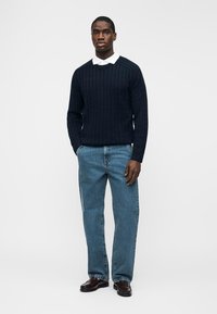 Uomo in piedi con le mani nelle tasche, indossando un maglione a trecce blu navy, una camicia bianca con colletto, jeans blu e scarpe marrone scuro.