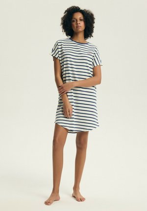 Kobieta stojąca boso, ubrana w białą sukienkę z krótkim rękawem w granatowe poziome paski, trzymająca dłonie z przodu.