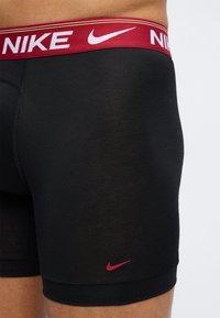 Κοντινό πλάνο ενός ατόμου που φοράει μαύρα μποξεράκια Nike με κόκκινη ζώνη μέσης και κόκκινο λογότυπο Nike στο αριστερό πόδι.