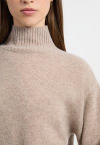 Suéter de cuello alto de punto grueso en color beige, con un cuello acanalado y una textura suave. Presenta mangas anchas y un corte holgado.