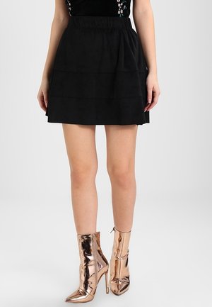 Woman wearing a black mini skirt and metallic rose gold high-heeled ankle boots, standing against a plain background.
