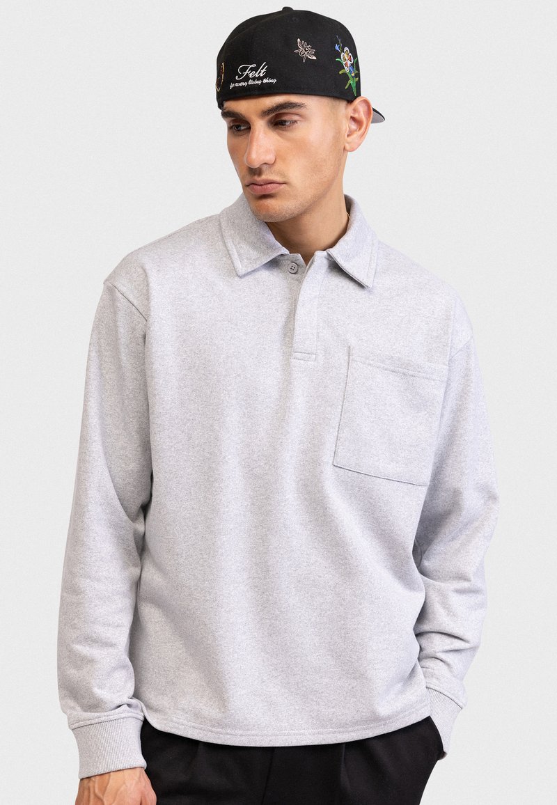 Ανοιχτό γκρι πόλο με μακριά μανίκια και γιακά, διακοσμημένο με τσέπη στο στήθος και ριμπ μανσέτες. Φοράει μαύρο καπέλο με κεντητές λεπτομέρειες.