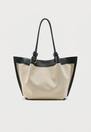 Μπεζ τσάντα ώμου από καμβά με μαύρες δερμάτινες λεπτομέρειες, χερούλια δεμένα σε κόμπο και μεταλλικά στοιχεία σε χρυσή απόχρωση, στέκεται όρθια πάνω σε απλό φόντο.