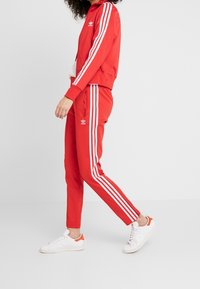 Röd track jacka och byxor tillverkade av slätt tyg, med vita tre-rands detaljer på ärmarna och sidorna. Vita sneakers kompletterar outfiten.