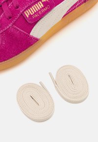 Baskets Puma Palermo en daim rose vif avec un accent en cuir blanc. Deux lacets plats en coton couleur crème sont présentés à côté de la chaussure.