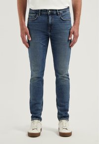 Kék farmernadrág, slim fit szabásban, öt zsebbel és tónusos varrással. Fehér fűzős sportcipővel párosítva.