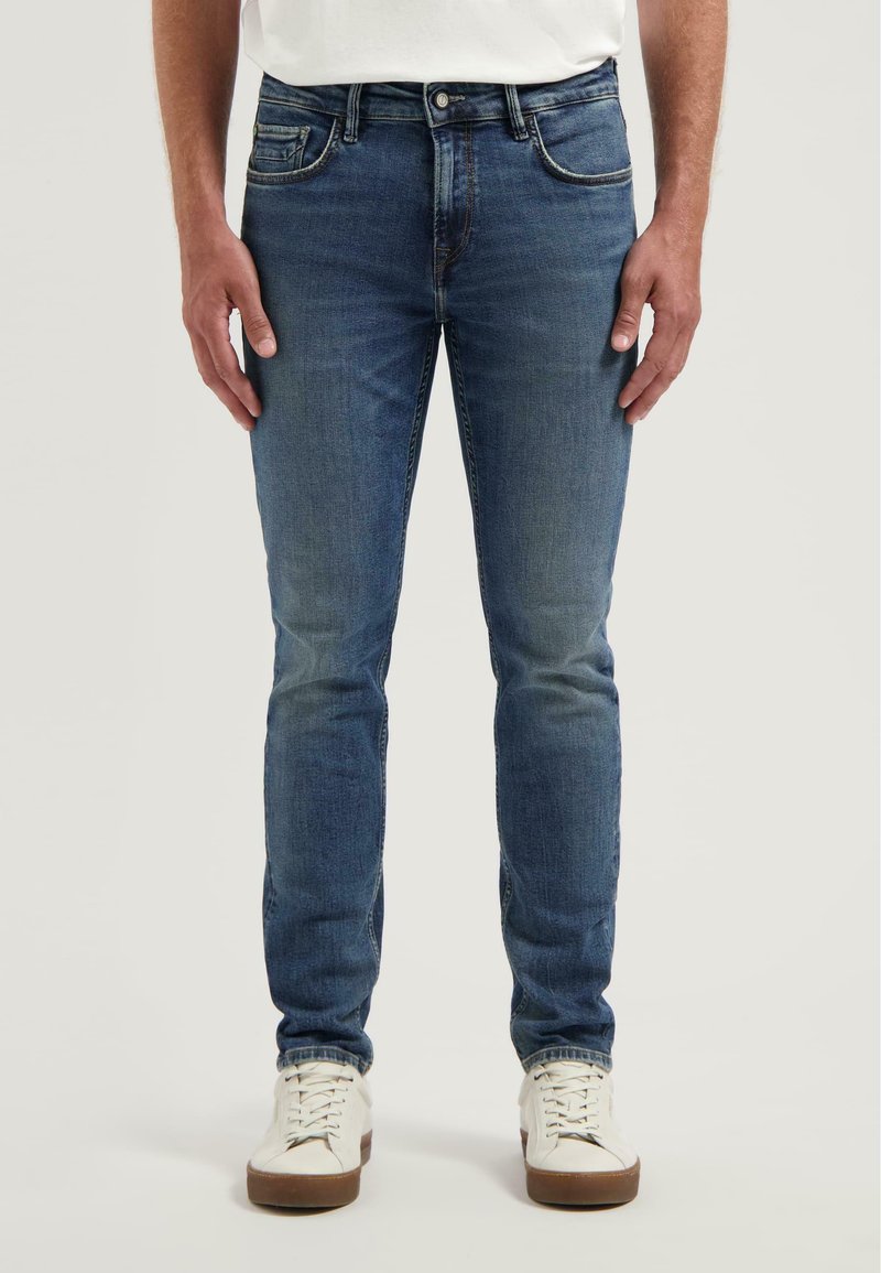 Kék farmernadrág, slim fit szabásban, öt zsebbel és tónusos varrással. Fehér fűzős sportcipővel párosítva.