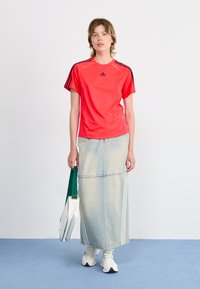 Red athletic t-shirt with black stripes on sleeves, paired with a light blue denim maxi skirt. White sneakers and a green strap bag.