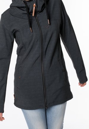 Vrouw draagt een zwarte zip-up hoodie met patroon, voorzakken en lichtblauwe spijkerbroek, tegen een witte achtergrond.