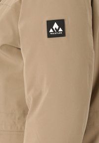 Vaalean beige päällystakki, joka on sileä ja jossa on musta neliölogo, jossa on tyylitelty vuoristomuotoilu hihassa.