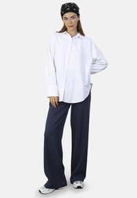 Camisa blanca de botones con un corte holgado, combinada con pantalones de pierna ancha azul oscuro y zapatillas grises. Una diadema negra con un diseño estampado completa el look.