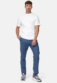Camiseta blanca de manga corta, pantalones azules, zapatillas grises con acentos rojos y azules. Tejido suave, corte casual, pose de pie, manos en los bolsillos.