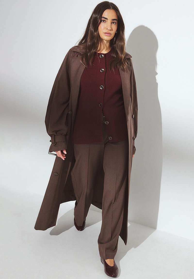 Brun trenchcoat över en vinröd button-up tröja och matchande byxor. Outfiten har mjuka texturer och en avslappnad passform, med spetsiga bruna skor.