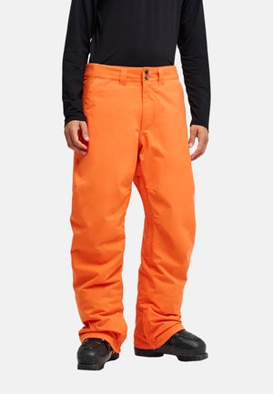 Mann trägt leuchtend orangefarbene Schneehose und schwarze Winterstiefel mit einem schwarzen langärmeligen Oberteil vor einfachem Hintergrund.