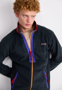 Čierna fleece bunda s oranžovým zipsom, fialovými akcentmi pozdĺž zipsu a na vreckách, červeným podšívka goliera a malým logom na hrudi.