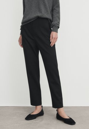 Person wearing black tapered pants, dark gray long-sleeve sweater, and black ballet flats standing on a gray floor against a white wall.