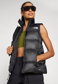 Svart pufferväst med dragkedjefickor och hög krage. Har en texturerad finish och North Face-loggan på bröstet. Bärs över en grön topp.