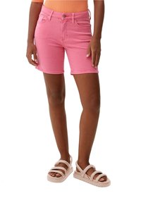 Persona vestida con shorts de mezclilla rosa, sandalias de tiras beige y una camiseta de canalé naranja, de pie con las manos relajadas a los lados.