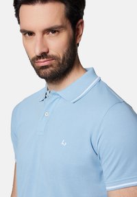 Jasnoniebieska koszulka polo z teksturowanej tkaniny i zapięciem na dwa guziki. Posiada białe logo na lewej piersi oraz białe paski na kołnierzyku.