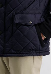 Navyblaue gesteppte Jacke mit Klapptasche und Druckknopf, beige Hose und eine Hand, die in eine Seitentasche gesteckt ist.