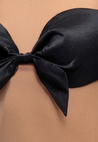 Czarny satynowy top bikini z wyraźną kokardą na środku, o gładkiej fakturze i konturowanym kształcie na nagim tle.