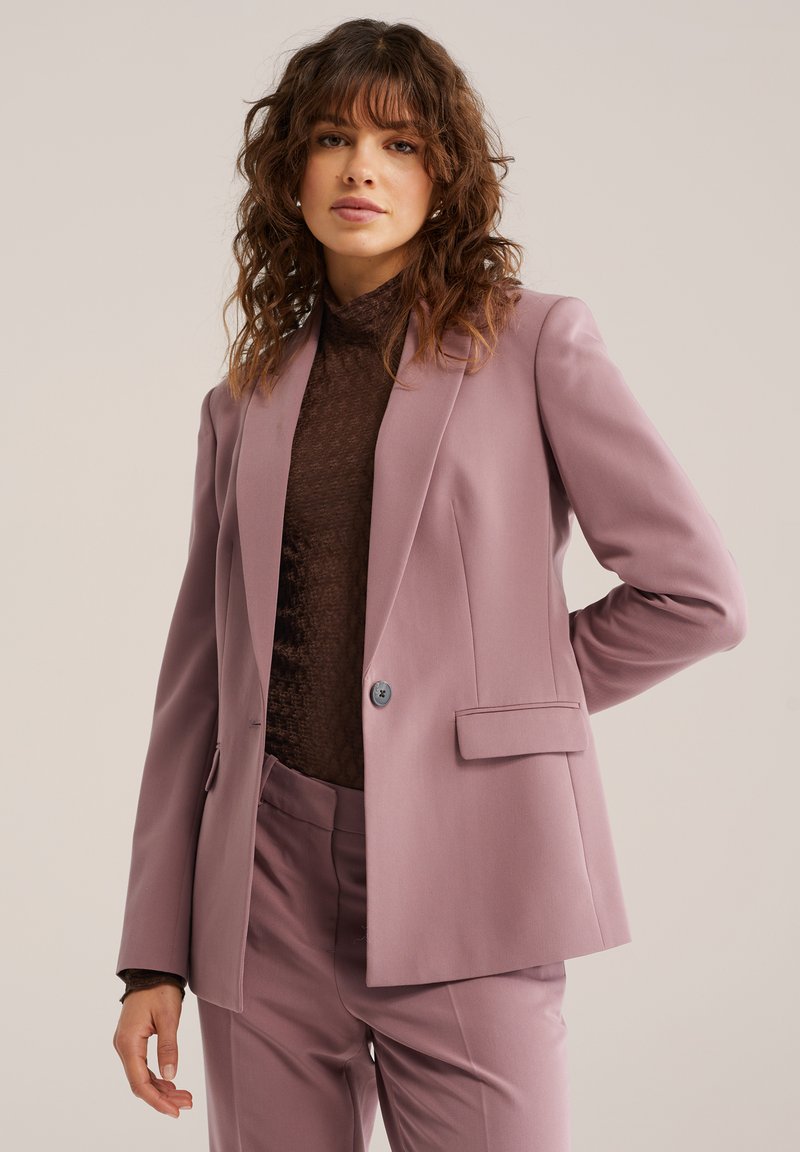 Svetlo vijoličast suknjič iz tkanine z gladko teksturo. Ima en gumb, zareze na reverjih in stranske žepe. Kombiniran z rjavim zgornjim delom.