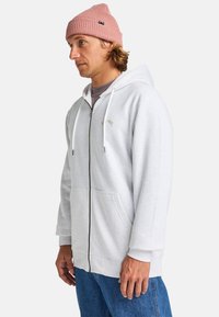 Sudadera con cremallera de color gris claro con dos bolsillos delanteros, cordones blancos y un pequeño logo dorado en el pecho. Combinada con un gorro de lana rosa.