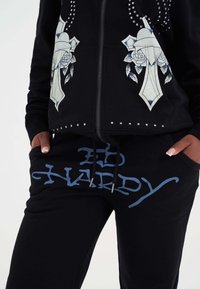 Felpa nera con zip e borchie argentate, con grafiche floreali e di spade. Pantaloni della tuta neri con testo blu "Ed Hardy", coulisse e tasche.