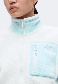 Άτομο που φοράει λευκό fleece μπουφάν με γαλάζιο fleece γιακά και τσέπη στο στήθος, με ορατό φερμουάρ και λογότυπο The North Face.