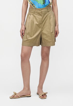 Kvinde iført lysebrune plisserede shorts, farverig top og beige sandaler med knyttede remme stående foran en ensfarvet hvid baggrund.